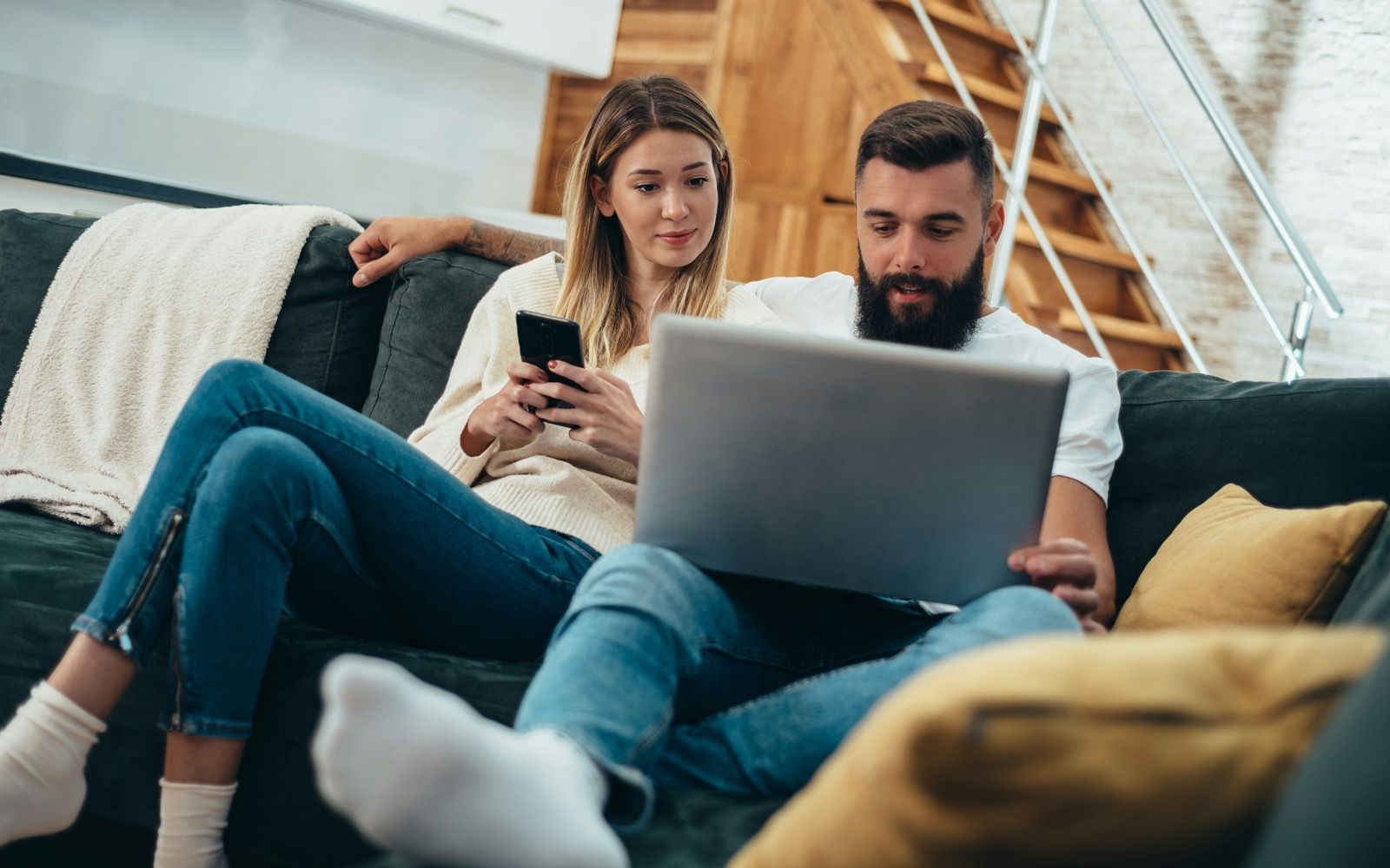 A couple relaxing on their couch at home, completing a quick health quiz on a phone and laptop